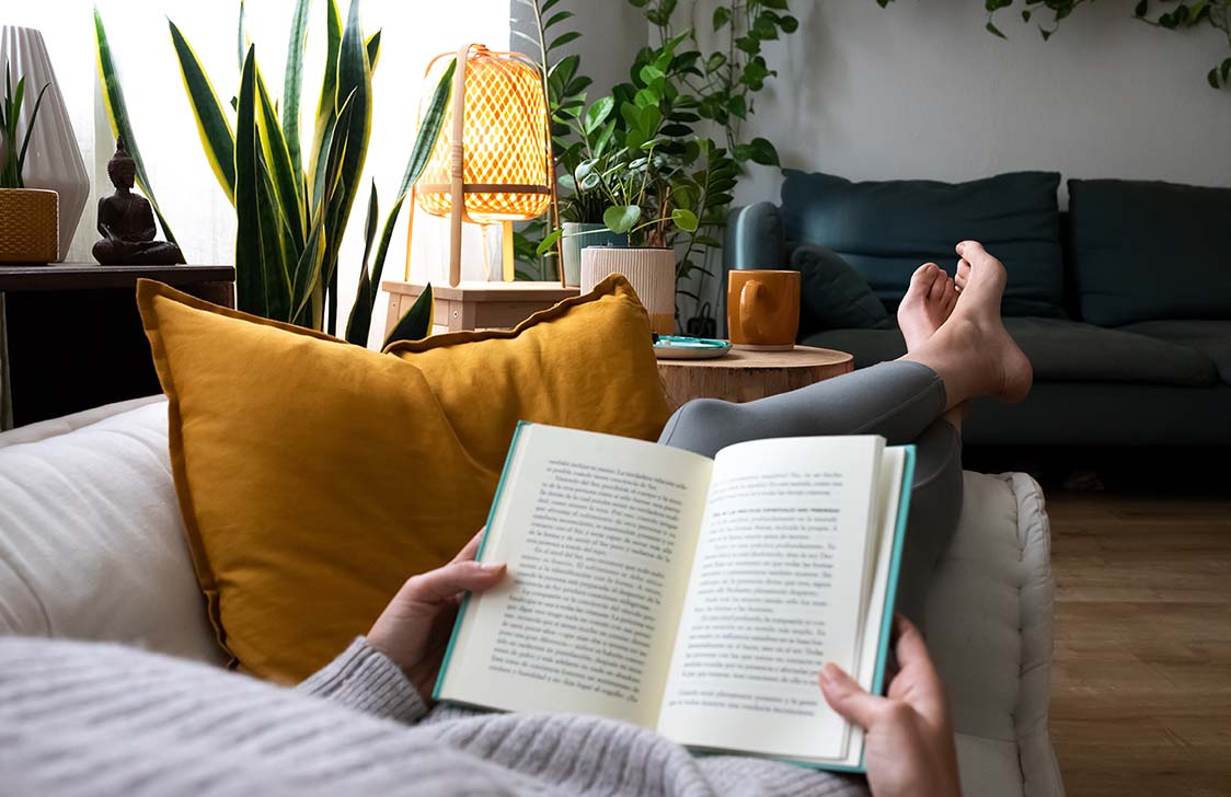 Jeune femme en train de se détendre à la maison, allongée sur un canapé et lisant un bon livre. 