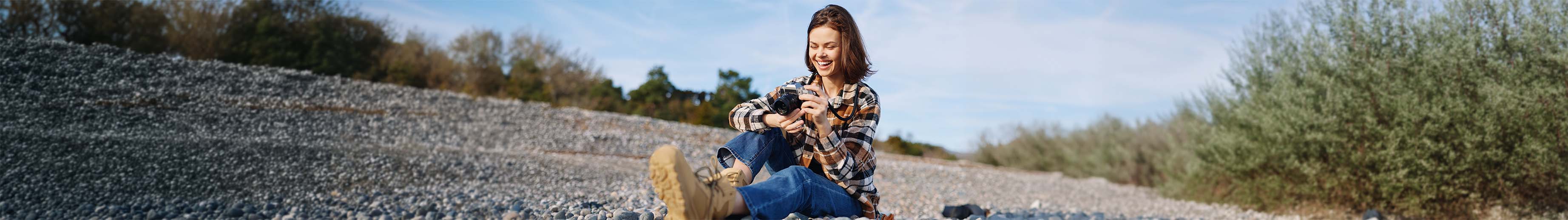 Personne assise sur une plage de galets tenant un appareil photo dans ses mains, vêtue d’une chemise à carreaux, d’un jean et de bottes de randonnée, avec des arbres et un ciel dégagé en arrière-plan.