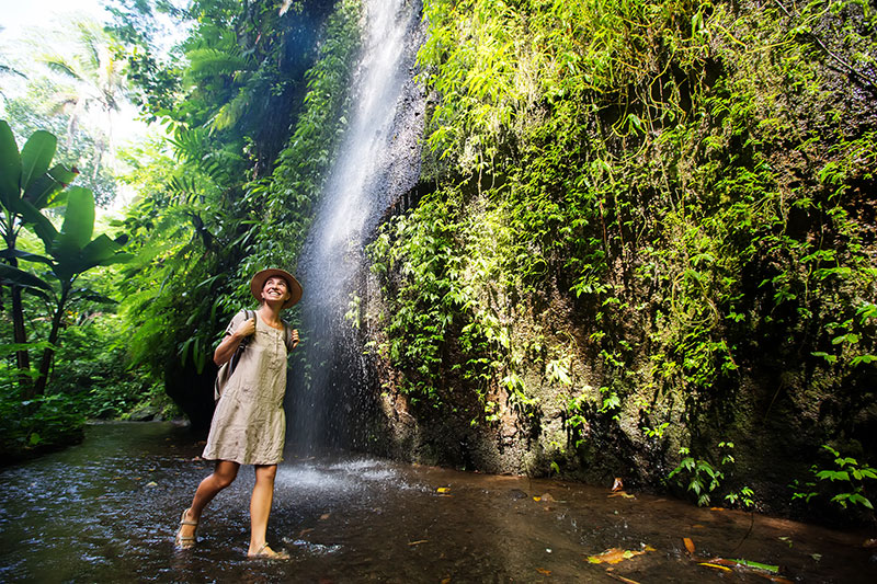 Une personne marchant dans un ruisseau peu profond sous une cascade, entourée de végétation luxuriante.